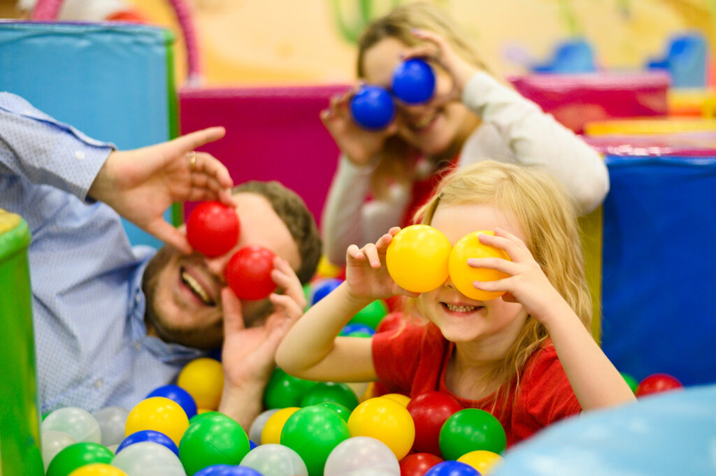 A family putting colourful plastic balls on their faces whilst sat in a soft play ball pit